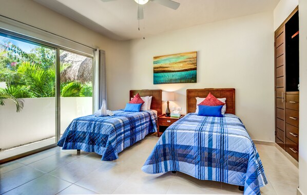 Habitación con dos camas, ventilador de techo, un cuadro y vista al verdor exterior.