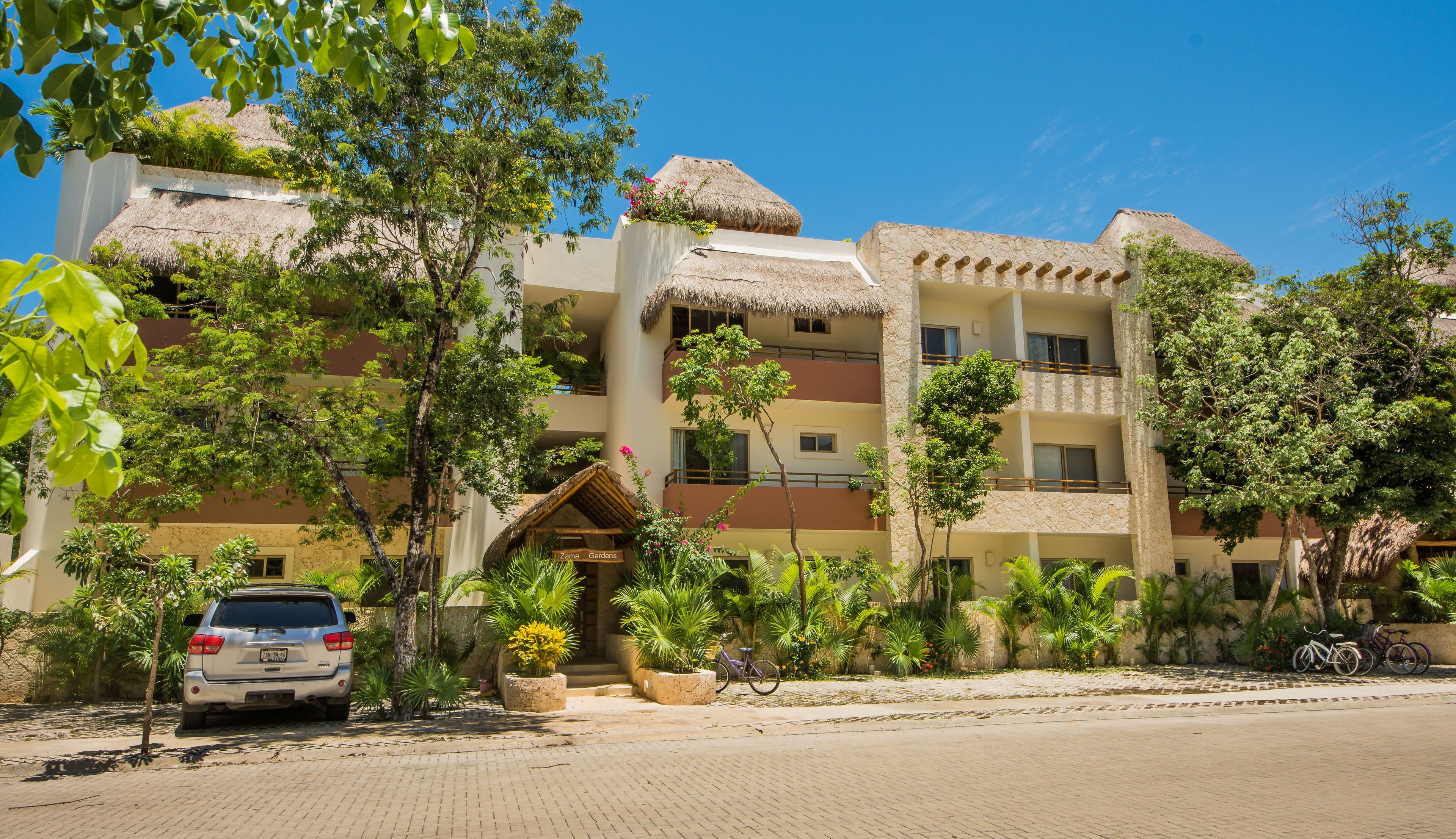 A multi-story hotel with a thatched roof, surrounded by lush greenery and a paved walkway.