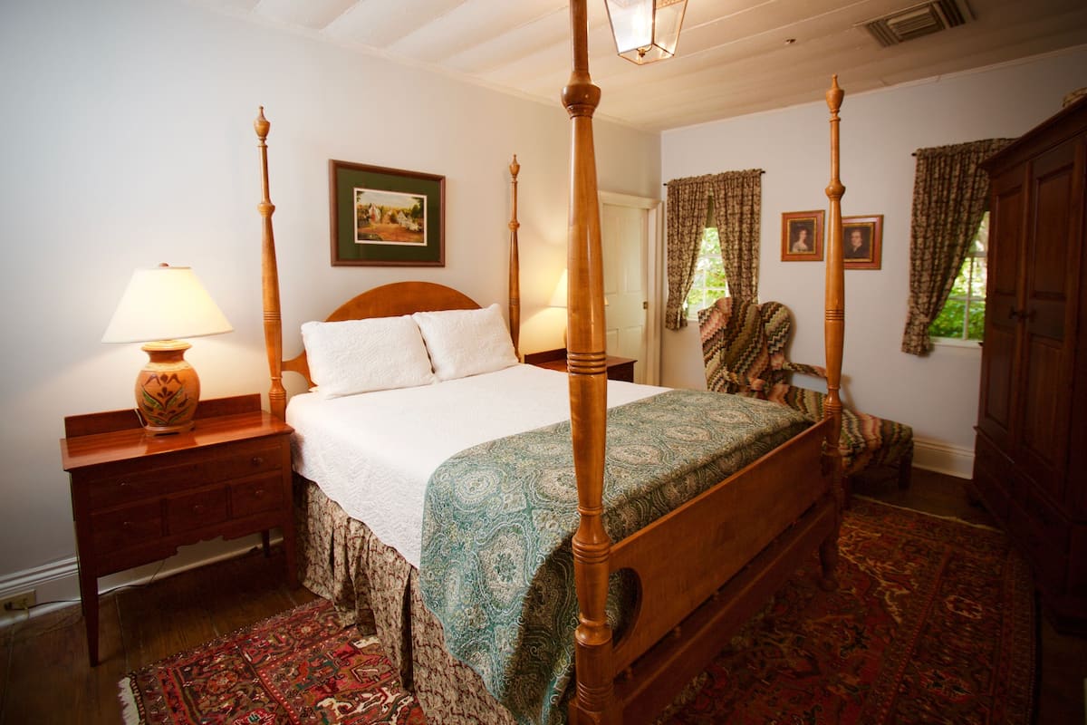 A four-poster bed with a wooden headboard, a bedside table with a lamp, a patterned rug, and a framed picture on the wall.