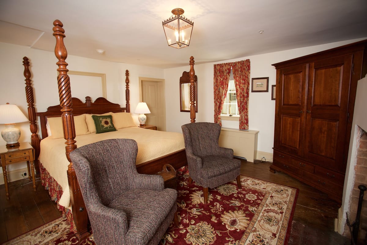 A bedroom with a four-poster bed, two armchairs, a wooden wardrobe, and a large window with curtains.