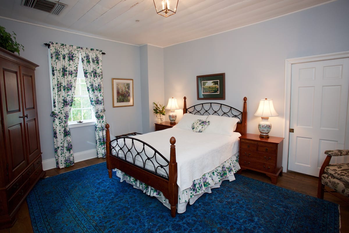 A bedroom with a wooden bed, floral curtains, a blue carpet, a wooden nightstand with a lamp, and a chair.