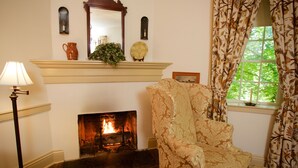 A cozy room with a fireplace, a patterned armchair, a lamp, and a window with curtains. - The Zevely Inn (Winston-Salem)