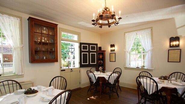 A dining room with wooden floors, white walls, and a chandelier. There are four dining tables with chairs, and a cabinet with glass doors. - The Zevely Inn (Winston-Salem)