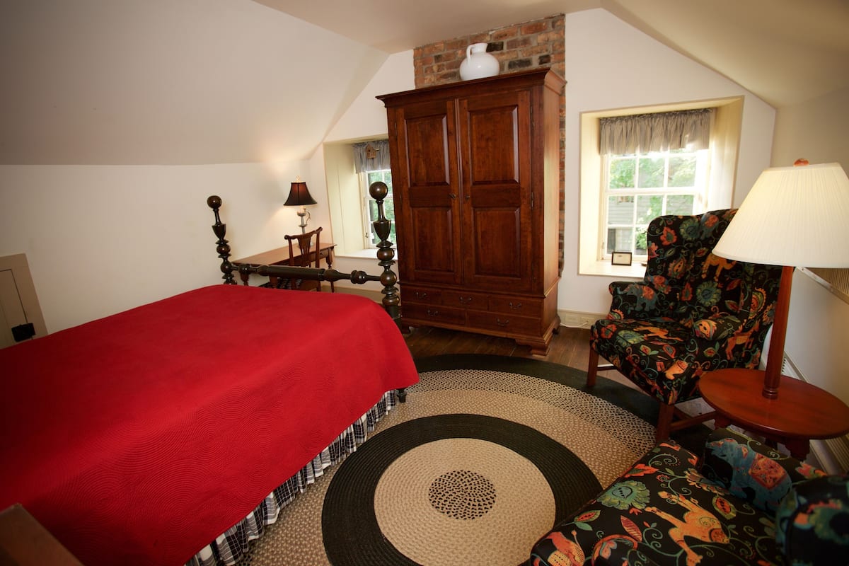 A bedroom with a bed, a wooden wardrobe, a chair, a lamp, and a window.