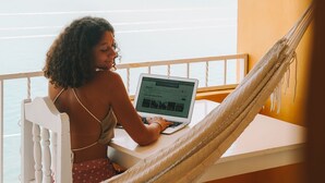 Uma pessoa usando um laptop numa varanda com vista para um corpo de água e colinas ao longe.