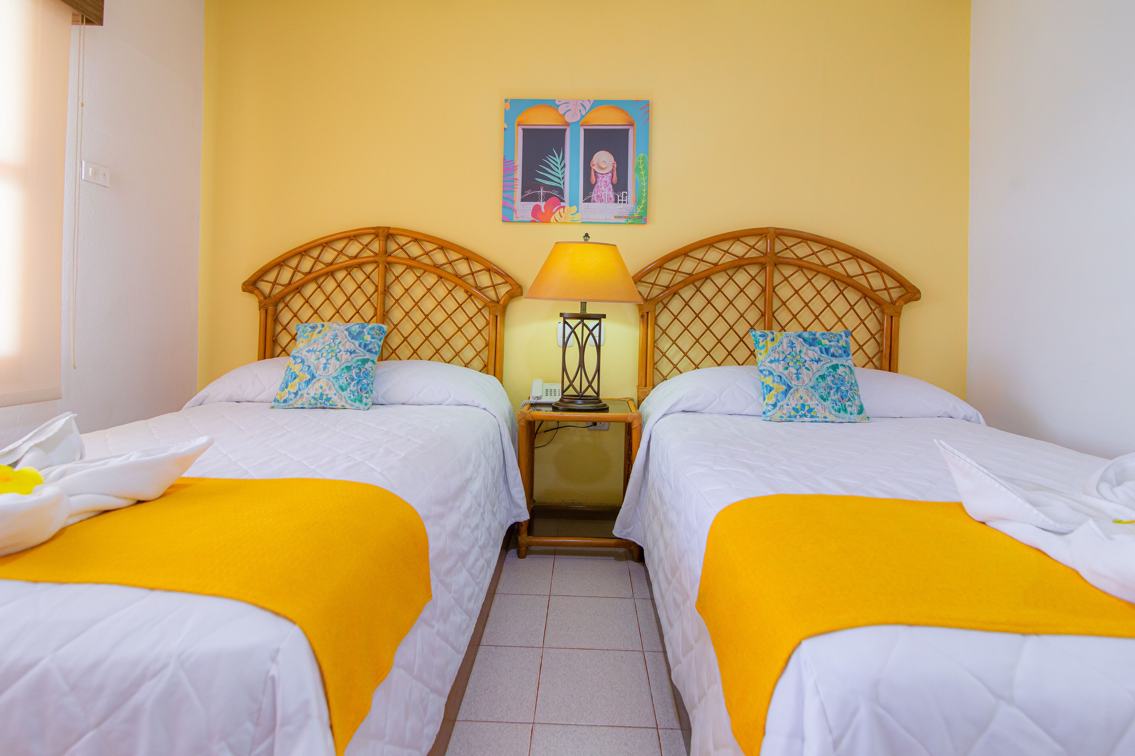 Habitación de hotel con dos camas, cada una con cubrecama amarillo y ropa de cama blanca. Hay una mesita de noche con lámpara y un cuadro sobre las camas.