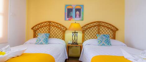Habitación de hotel con dos camas, cada una con cubrecama amarillo y ropa de cama blanca. Hay una mesita de noche con lámpara y un cuadro sobre las camas.