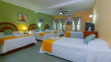 A hotel room with three beds, each with white bedding and orange accents. There is a ceiling fan, a television on a wooden table, and artwork on the walls.