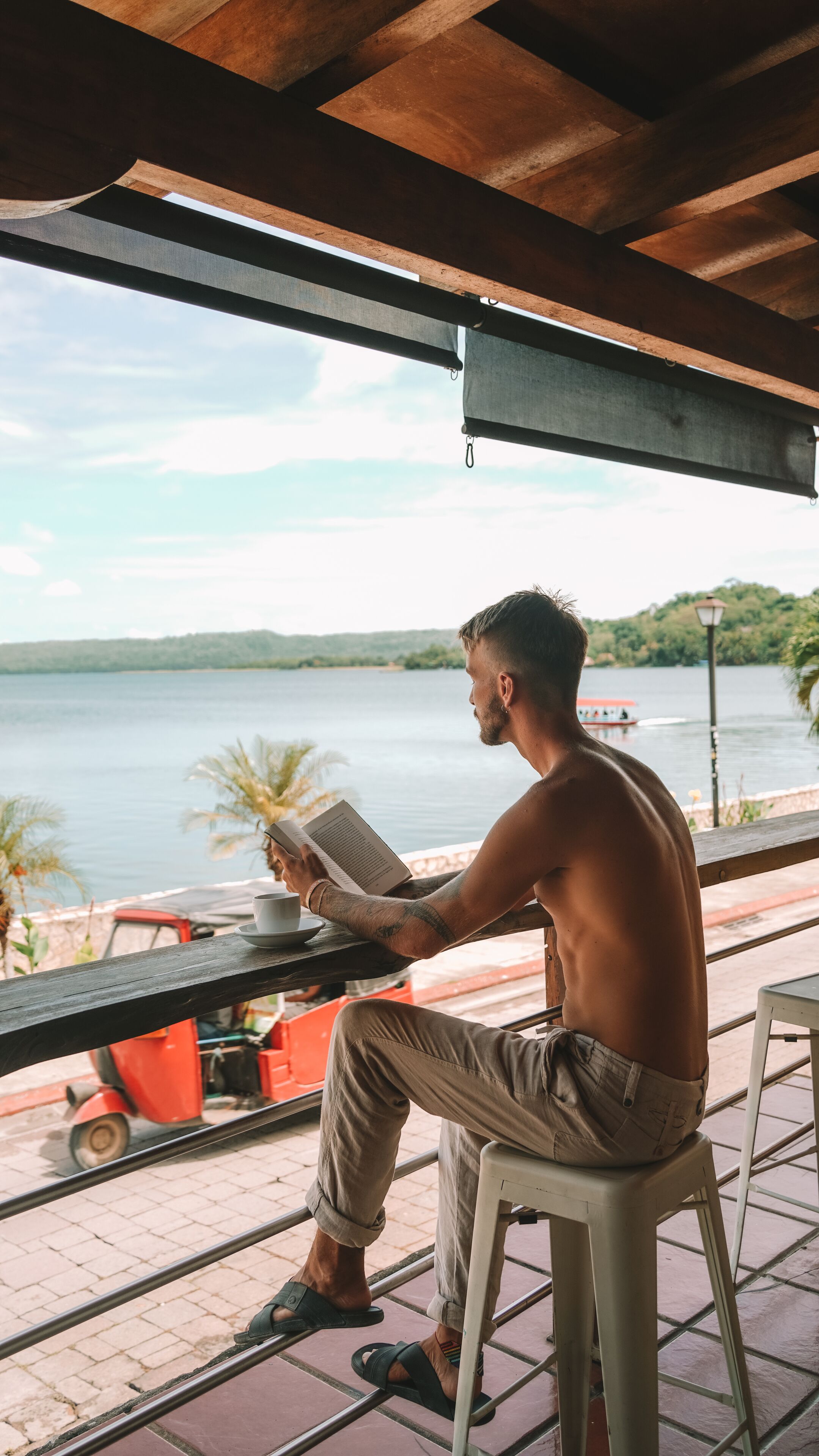A man reading a book under a wooden pergola with a view of a calm lake and a boat in the distance.