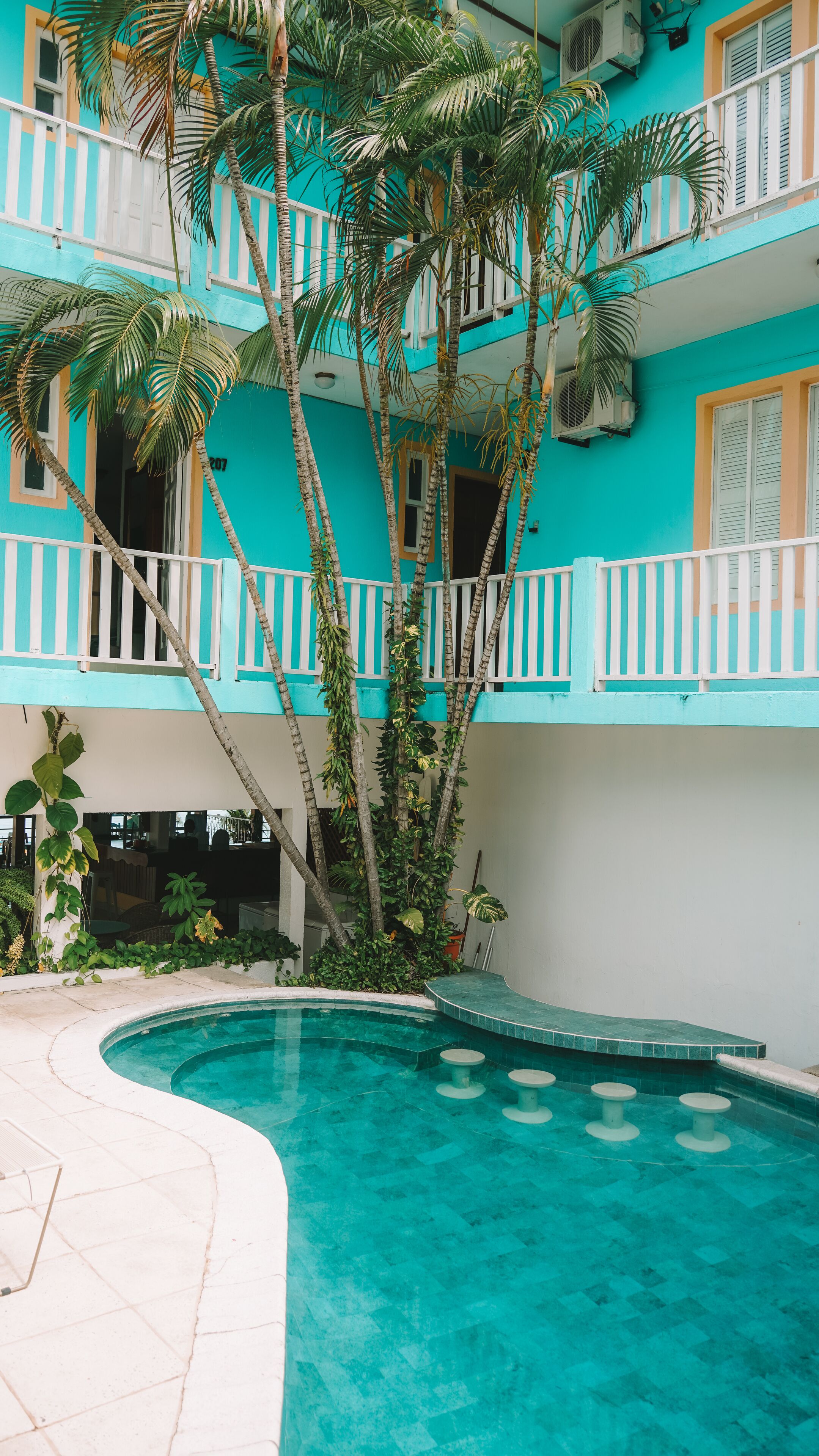 A two-story building with a pool, surrounded by palm trees and a white fence.