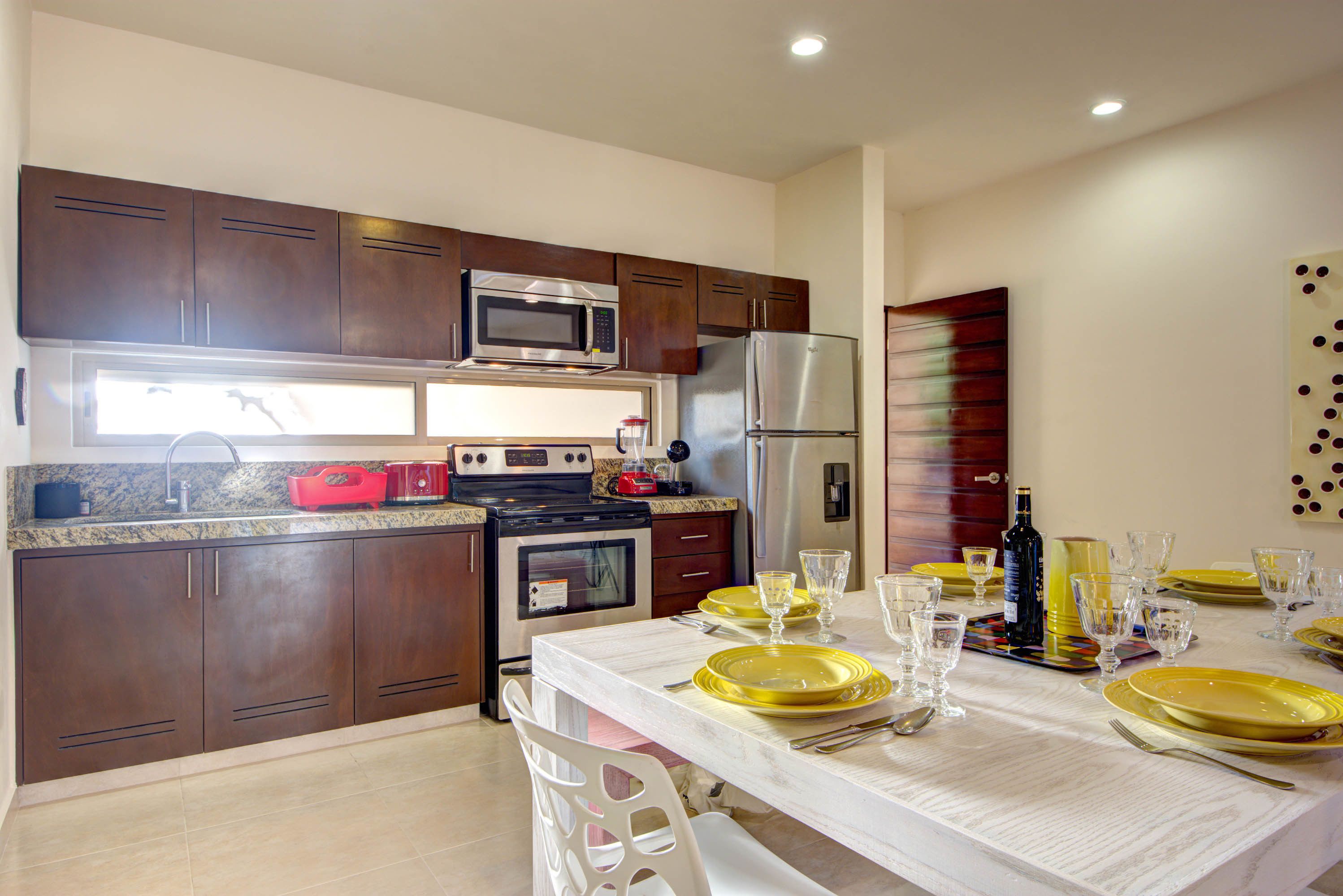 A modern kitchen with dark wooden cabinets, stainless steel appliances, and a dining table set with yellow plates and glasses.