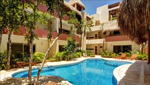 A swimming pool with wooden loungers and thatched umbrellas, surrounded by lush greenery and a multi-story building with balconies. - Rainbow Penthouse 2 bedrooms sleeps 5 (Tulum)