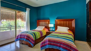 A room with two beds, each with a wooden headboard and colorful striped bedding. There is a ceiling fan, a window with a view of greenery, and a door to the right. - Rainbow Penthouse 2 bedrooms sleeps 5 (Tulum)
