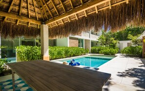 Un área de piscina con una mesa de madera, una persona en el agua y una estructura cubierta de paja.