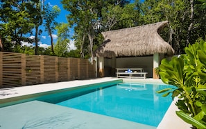 Una piscina con una estructura de techo de paja, rodeada de exuberante vegetación y una valla de madera.
