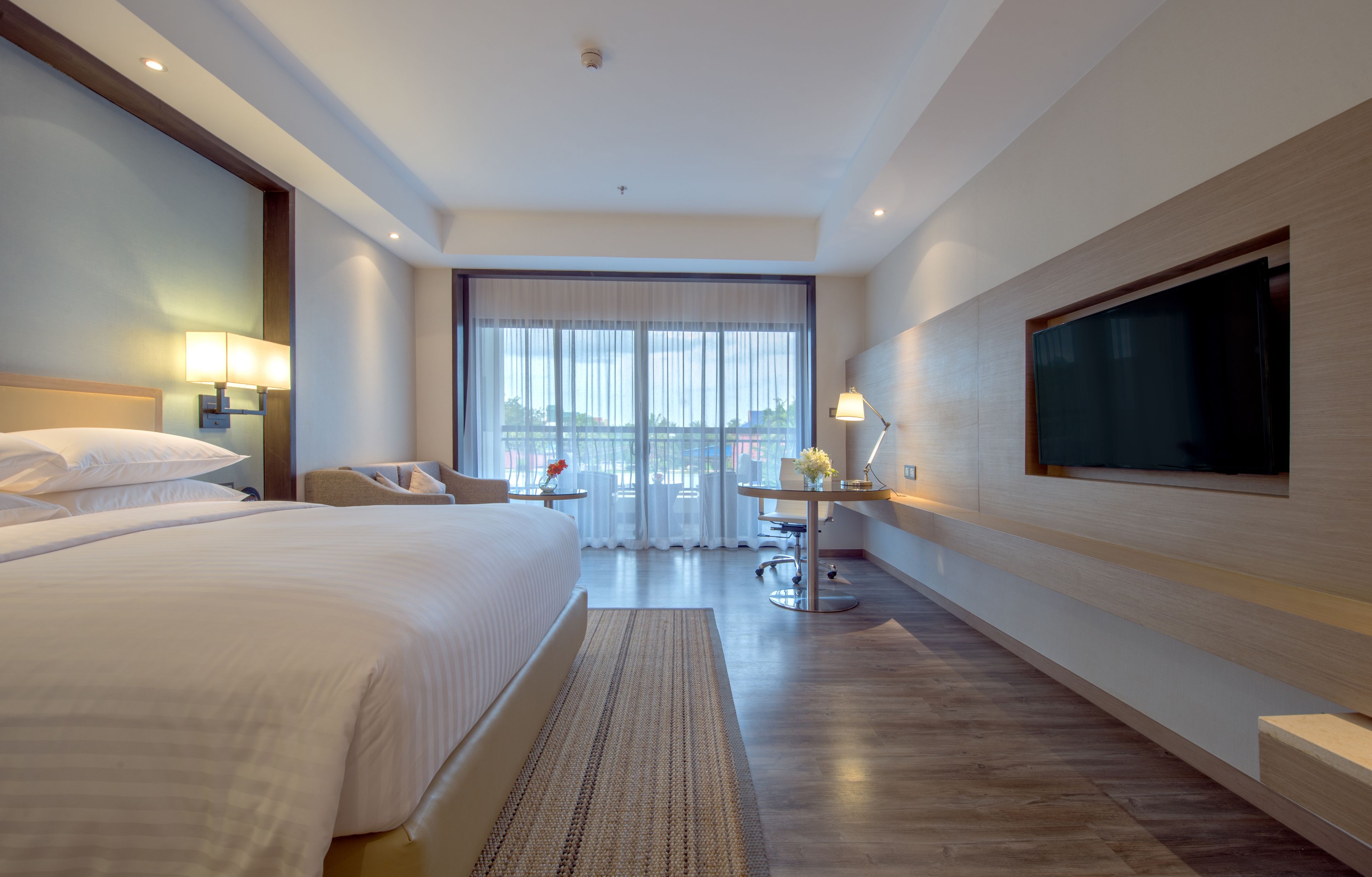 A modern hotel room with a large bed, a desk with a chair, a TV, and a view of the city through large windows.