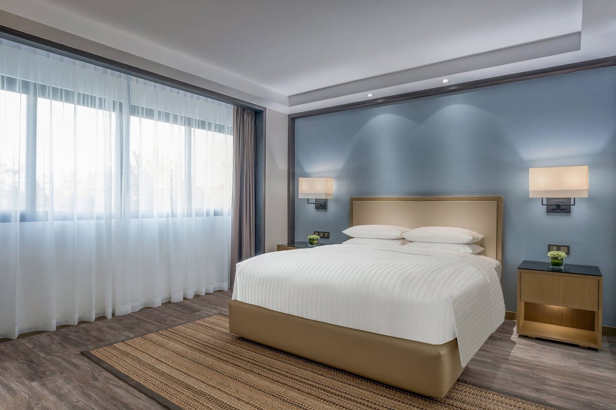 Une chambre d’hôtel moderne avec un grand lit, deux tables de chevet avec des lampes, une vue à travers des rideaux légers et un mur d’accent bleu.