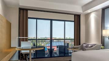 Una habitación de hotel moderna con un ventanal grande, una cama, un escritorio con silla y un pequeño balcón con mobiliario exterior.
