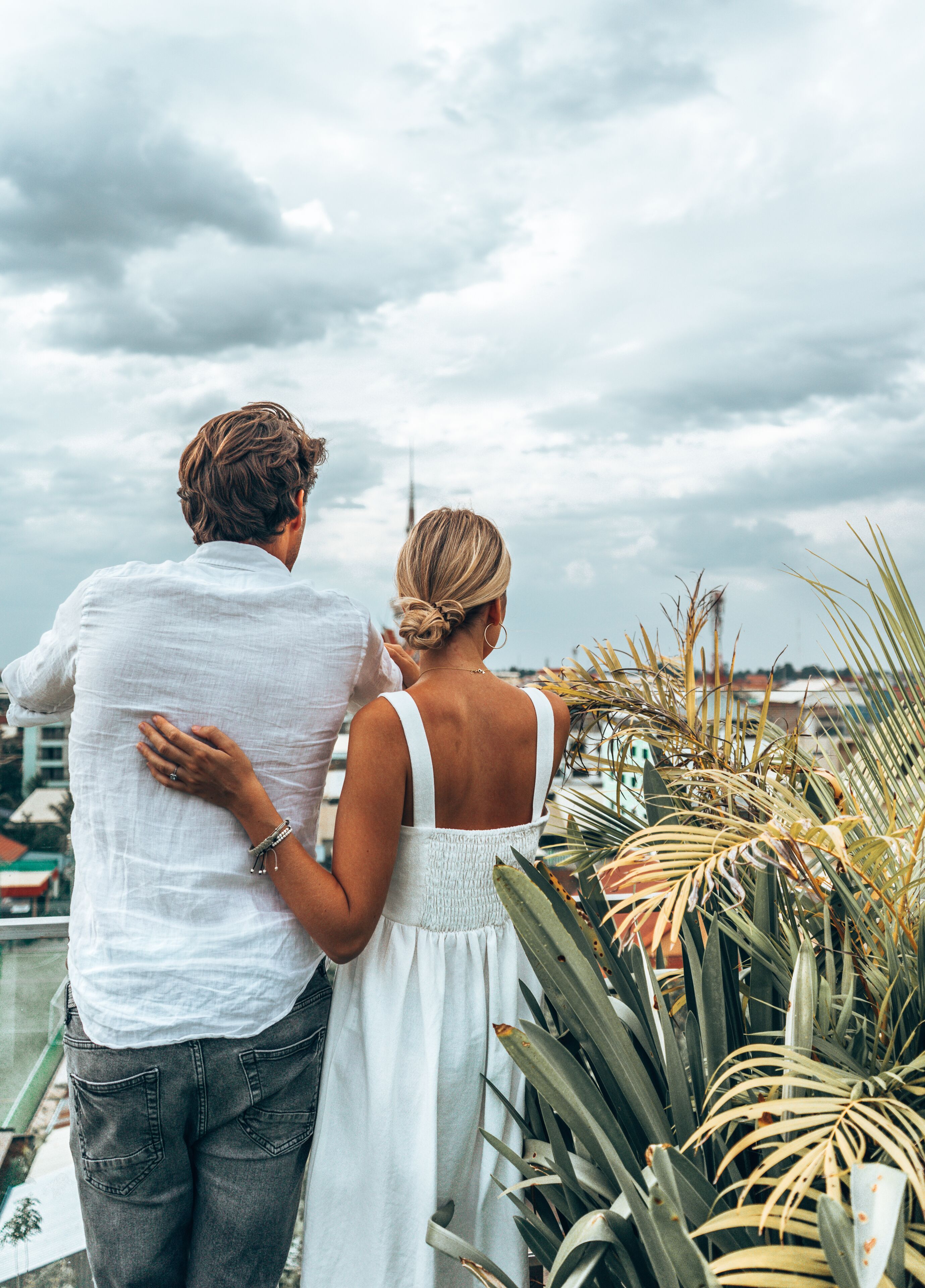 A couple standing on a balcony overlooking a cityscape with a cloudy sky.