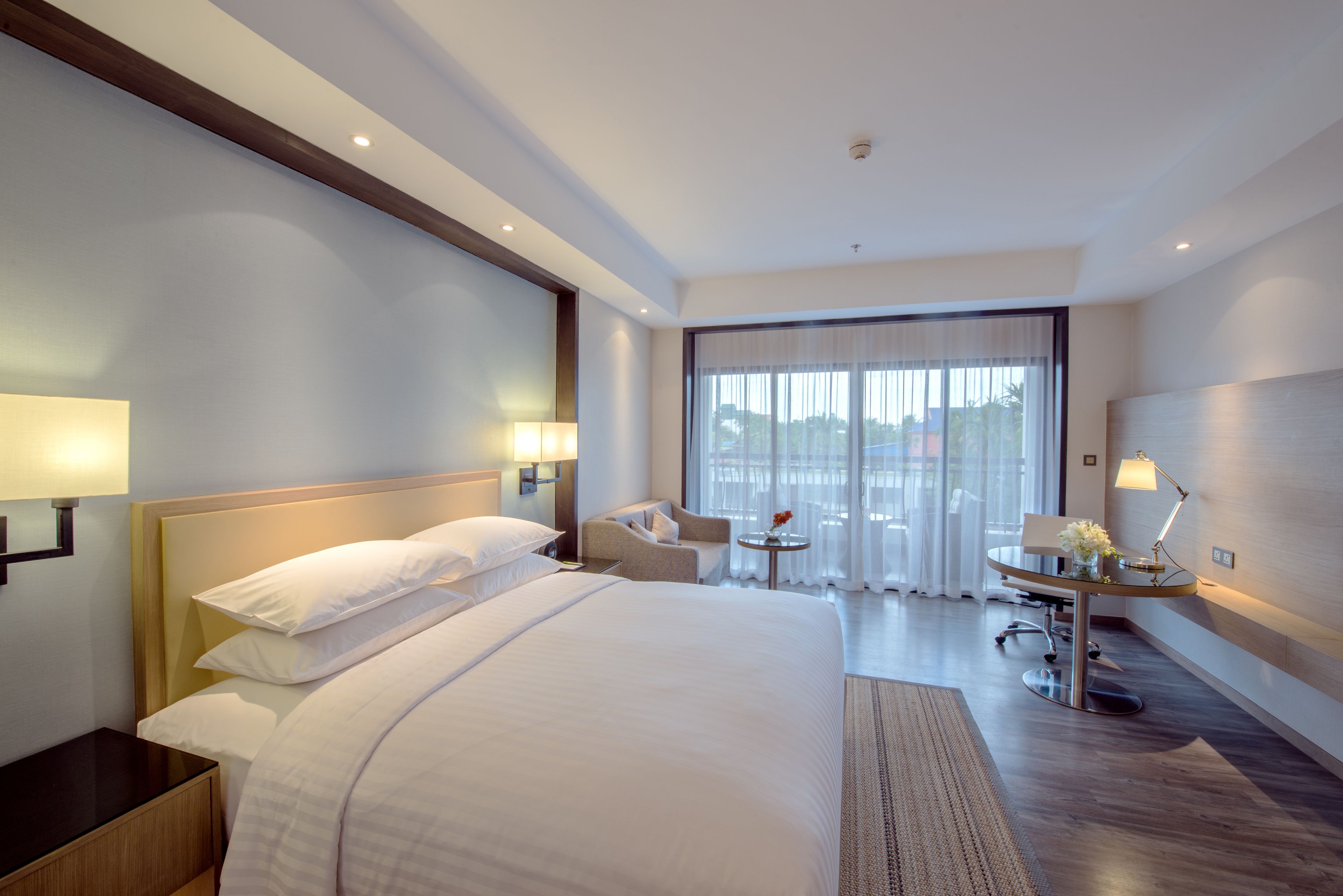 Ein Hotelzimmer mit einem großen Bett, einem Schreibtisch mit Stuhl, einem kleinen Tisch mit Lampe und Blick ins Freie durch große Fenster.