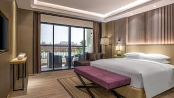 A modern hotel room with a large bed, a bench, a desk, and a balcony with outdoor furniture.