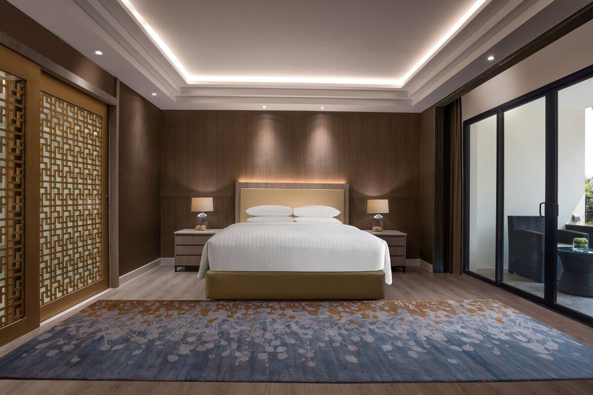 Une chambre d’hôtel moderne avec un grand lit, des murs recouverts de panneaux en bois et une vue sur l’extérieur, accessible par une porte-fenêtre coulissante.