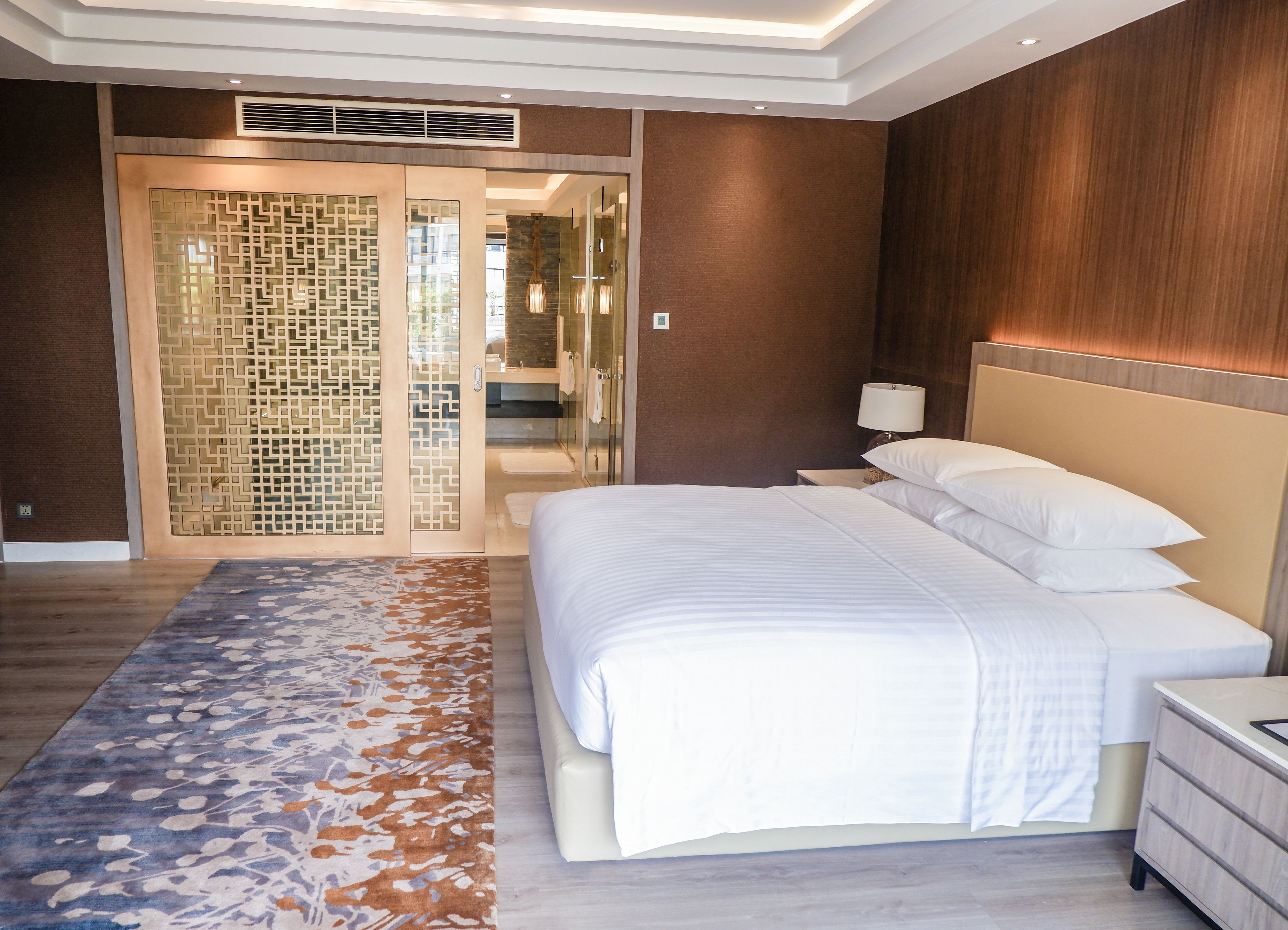 A modern hotel room with a large bed, a nightstand, a lamp, and a decorative wall panel.