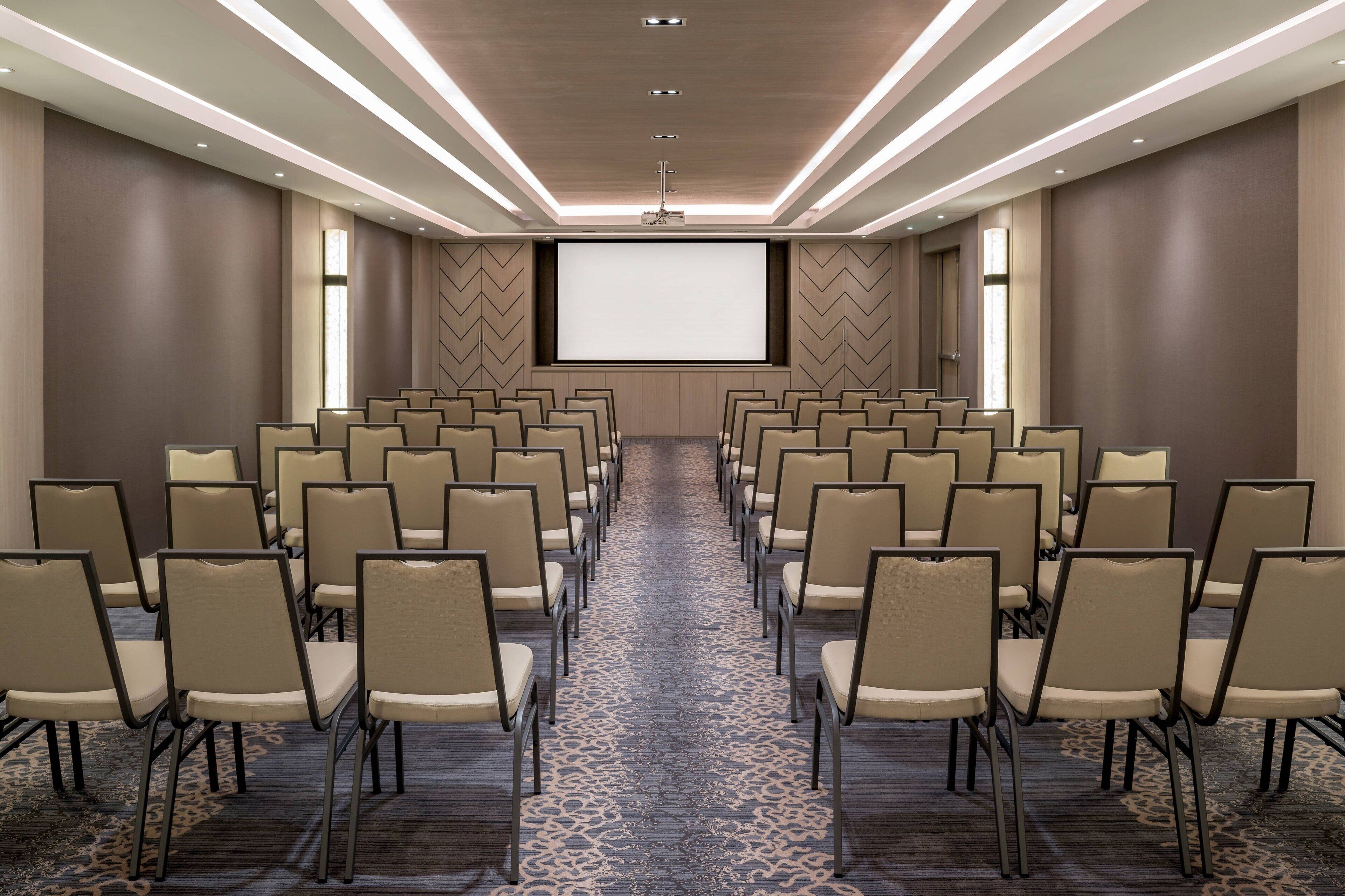 A conference room with rows of chairs, a large screen, and a carpeted floor.