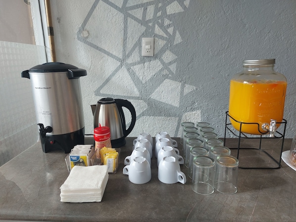 Una zona de cocina con un dispensador de agua caliente Hamilton Beach, una cafetera, una botella de condimentos, una pila de servilletas y una jarra grande de jugo de naranja.