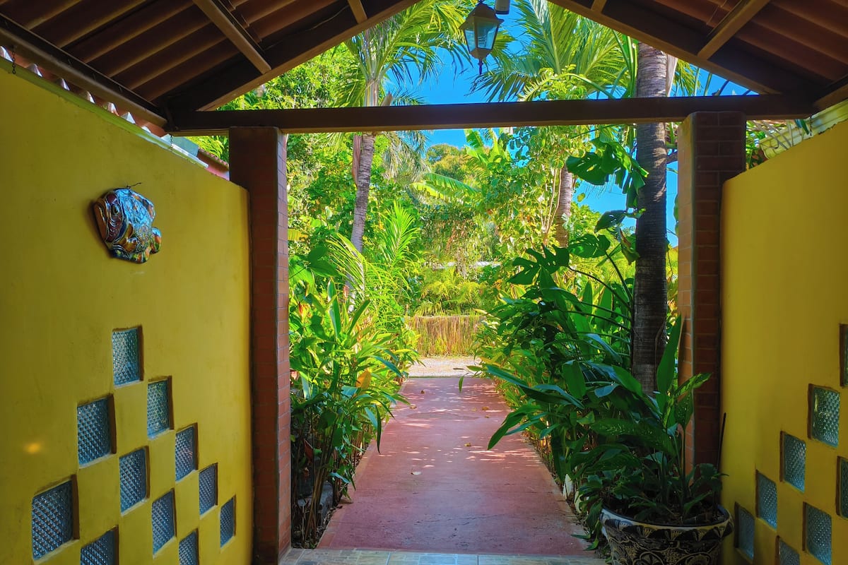 Un chemin couvert mène à un jardin luxuriant, dont le sol est pavé de briques rouges et les murs peints en jaune, et orné de carreaux décoratifs.