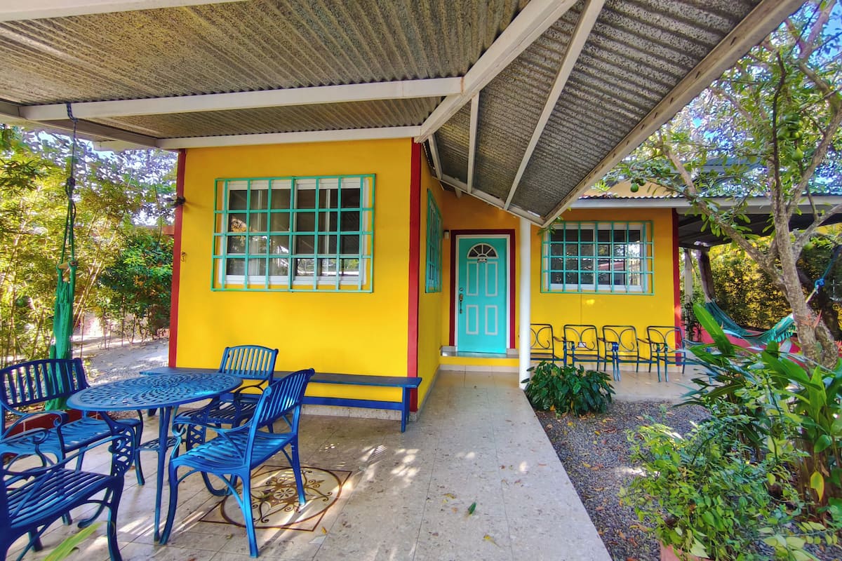 Un patio abrité avec un bâtiment jaune, des chaises bleues et une table.