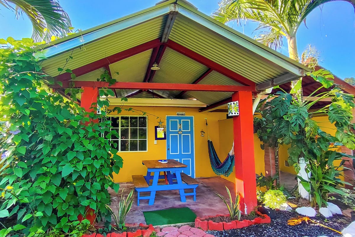 Un bâtiment jaune avec un toit rouge et une porte bleue. Un hamac est suspendu sur le côté.