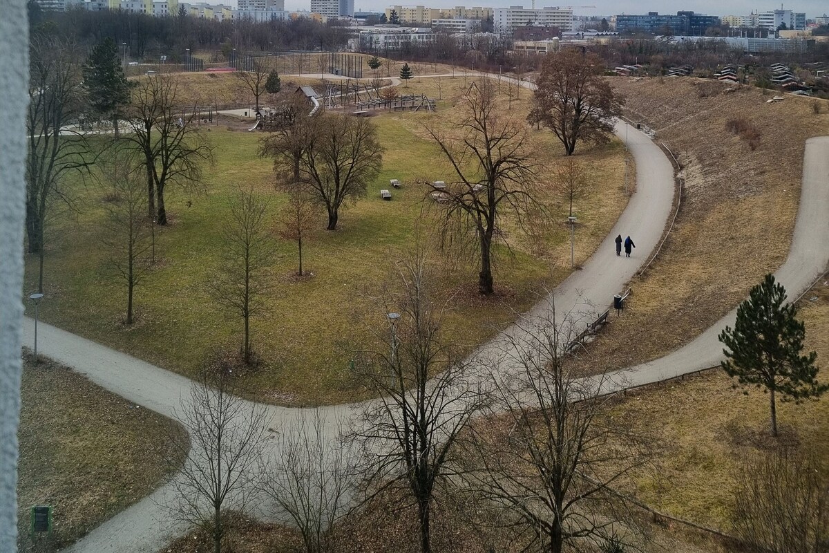Un parc avec des sentiers, des arbres et des bâtiments qui se dessinent à l’horizon sous un ciel nuageux.