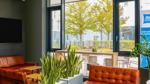 A modern indoor seating area with leather couches and tables, large windows with a view of trees and buildings, and a sign for 'ibis budget'. - Ibis Budget Muenchen City Olympiapark (Munich)