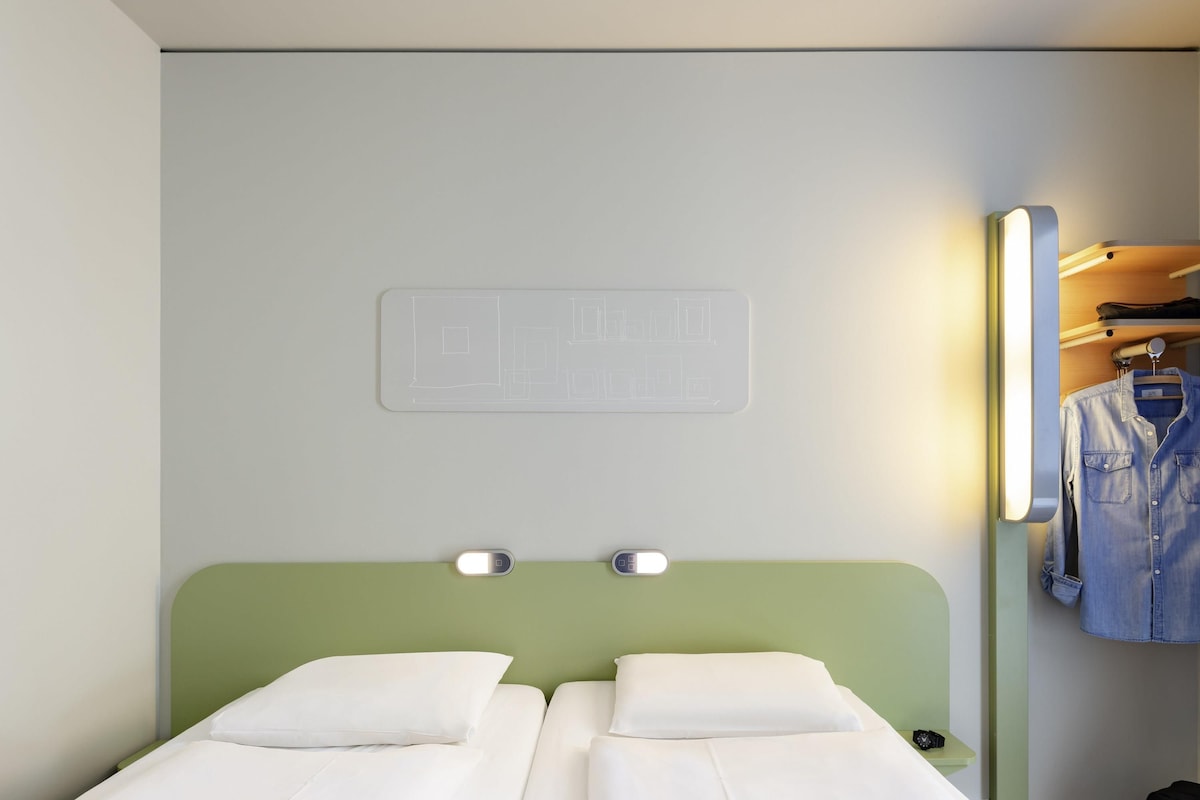 A hotel room with two beds, a green headboard, a wall-mounted light, a mirror, and a clothing rack.