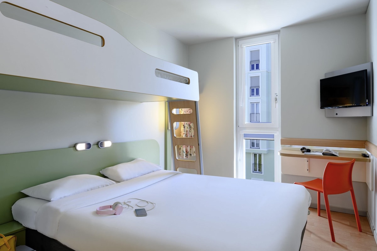 Une chambre d’hôtel équipée d’un lit, d’un bureau, d’une chaise, d’une télévision et d’une fenêtre donnant sur des immeubles.