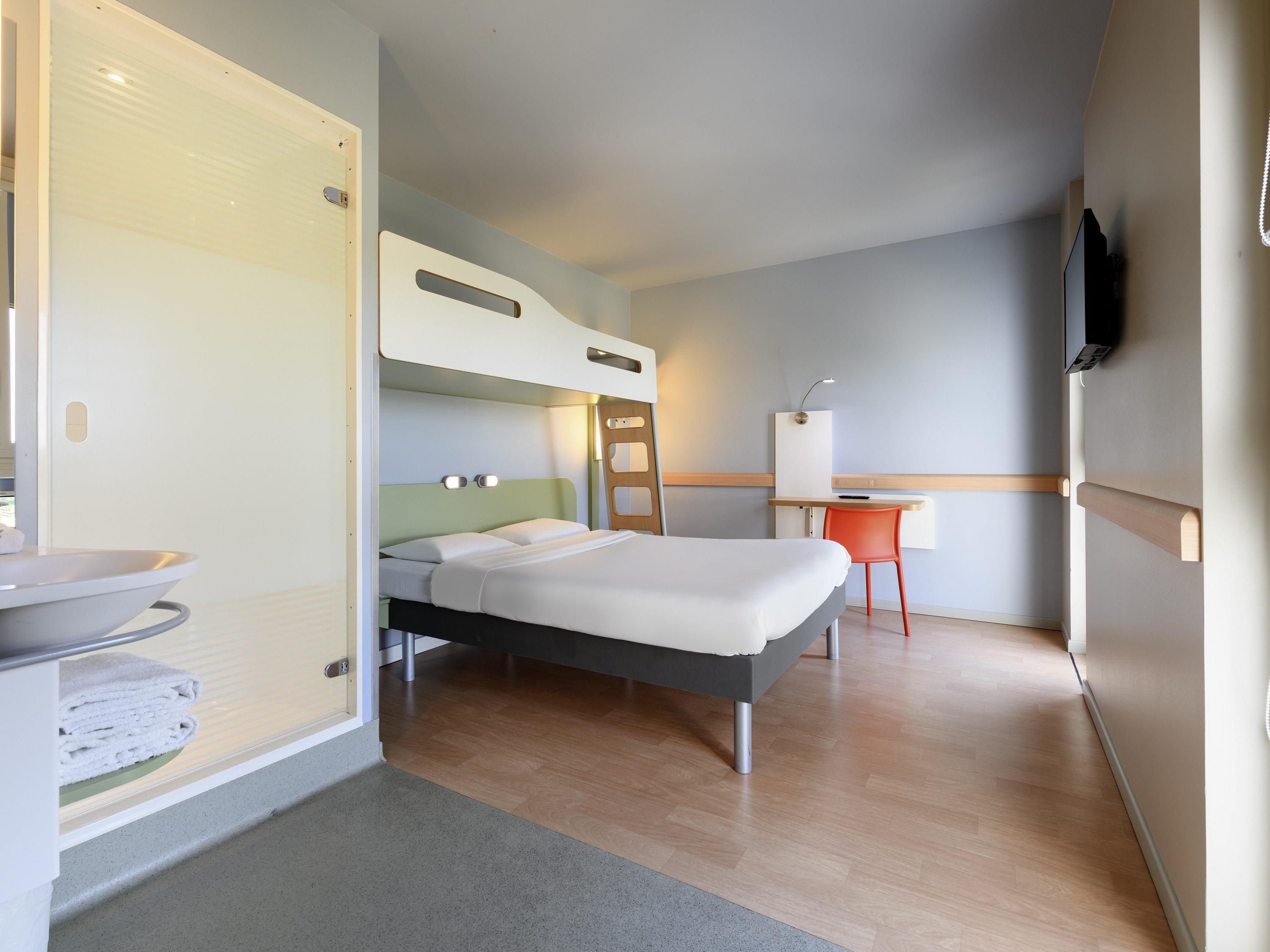 A hotel room with a bed, a desk, a chair, a TV, and a bathroom with a sink and towel rack.