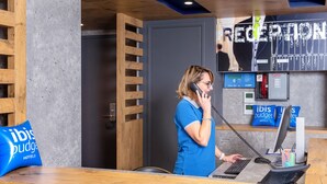 Un comptoir d’accueil en bois, un téléphone et un ordinateur se trouvent à la réception de l’hôtel. Des coussins bleus ornés du logo de l’hôtel sont disposés sur le comptoir, ainsi qu’une enseigne indiquant « RÉCEPTION ».