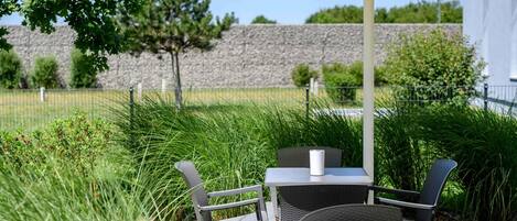 An outdoor patio with a table and chairs under a yellow umbrella, surrounded by greenery and a stone wall.