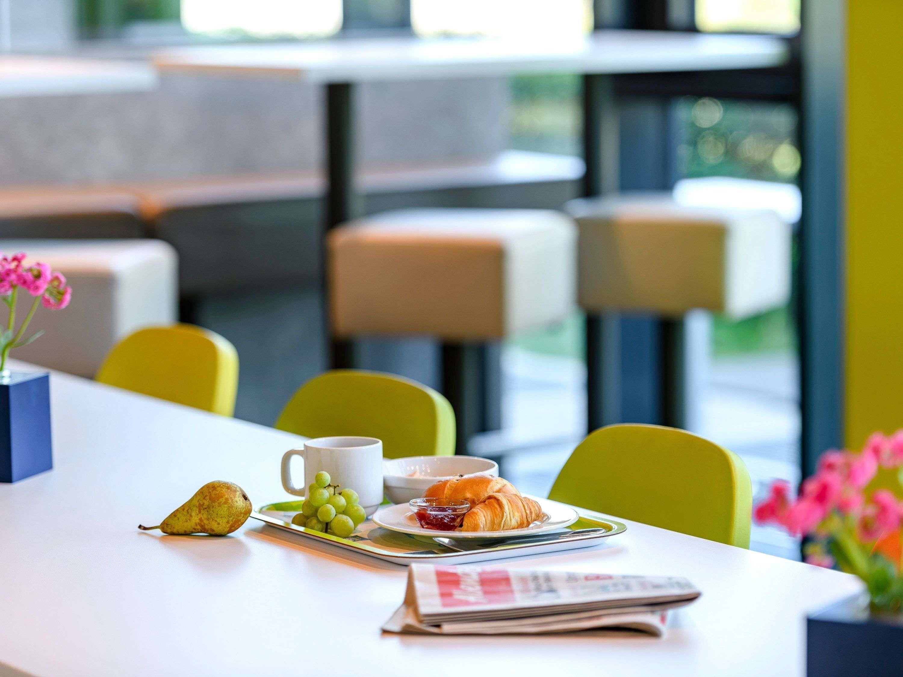 A dining table with a pear, grapes, a croissant, a cup, and a newspaper. There are yellow chairs and a vase with pink flowers.