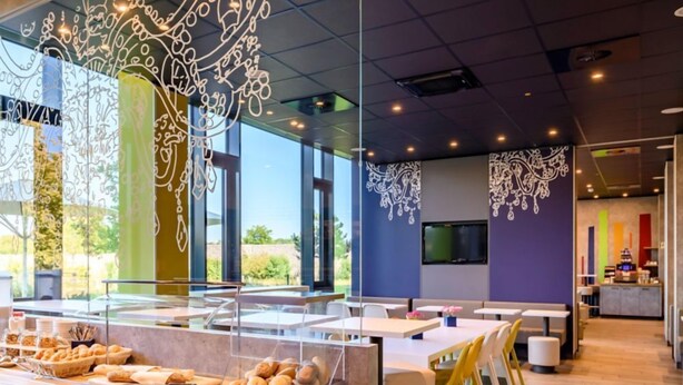 Un café moderne avec un comptoir central où sont exposées des pâtisseries, un espace salon avec des tables et des chaises, et des œuvres d’art décoratives sur les murs.