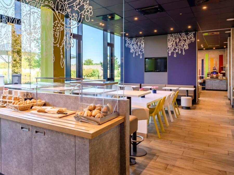 A modern cafe with a central counter displaying pastries, a seating area with tables and chairs, and decorative wall art.