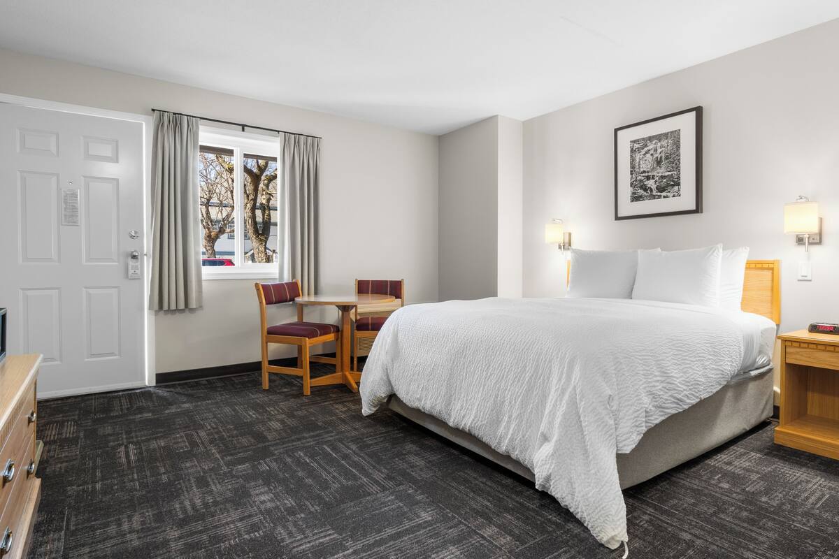 Ein Hotelzimmer mit Bett, Schreibtisch, Stuhl, einem fenster mit Vorhängen, einem gerahmten Bild an der Wand und einem Nachttisch mit Lampe.