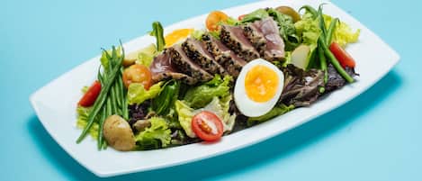 Een wit rechthoekig bord met een salade van sla, kerstomaatjes, groene bonen en in plakjes gesneden vlees, vergezeld van een gekookt ei in tweeën.