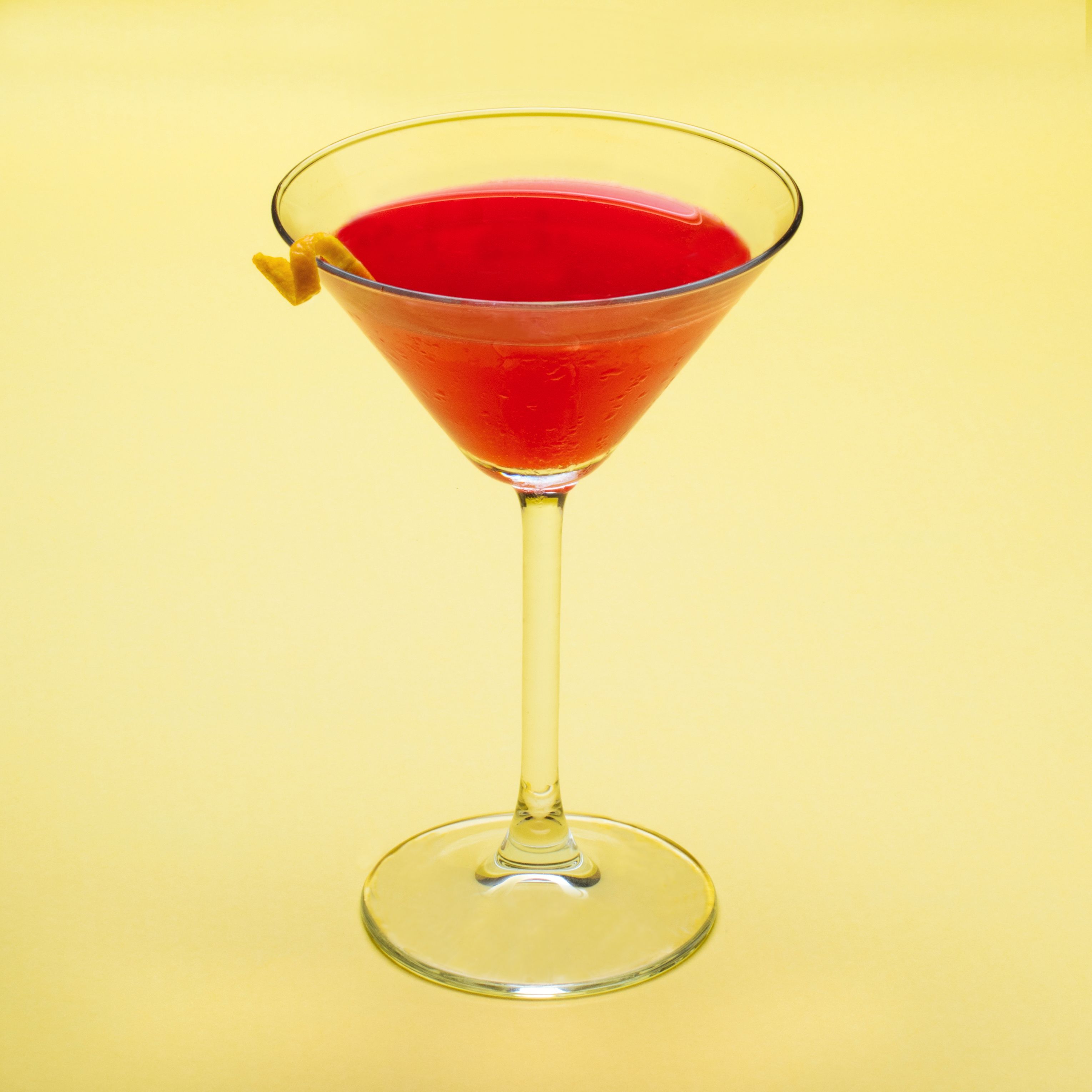 Una copa de martini con un líquido rojo y una aceituna de limón como decoración sobre un fondo amarillo.
