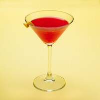 Una copa de martini con un líquido rojo y una aceituna de limón como decoración sobre un fondo amarillo.