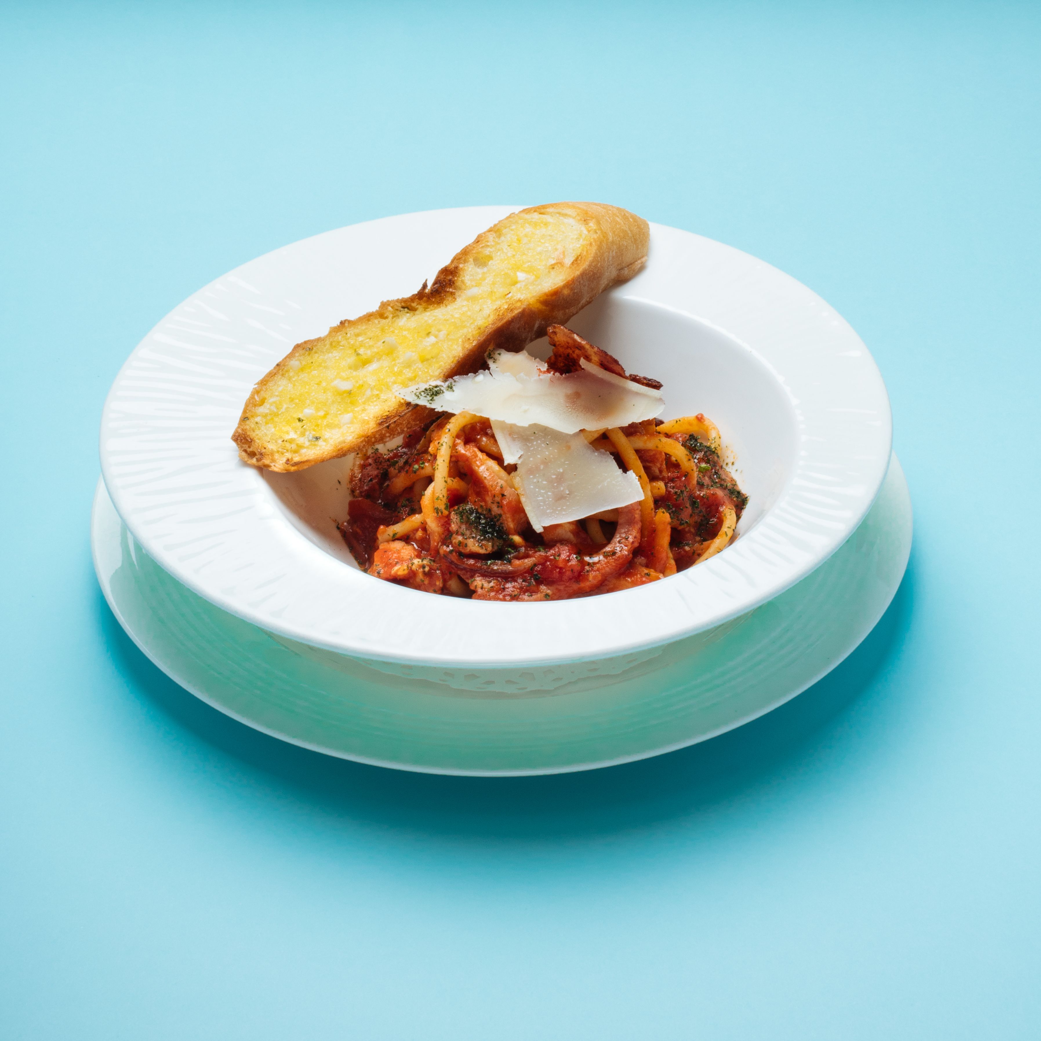 A white plate with a pasta dish, topped with a slice of bread and grated cheese, on a blue background.