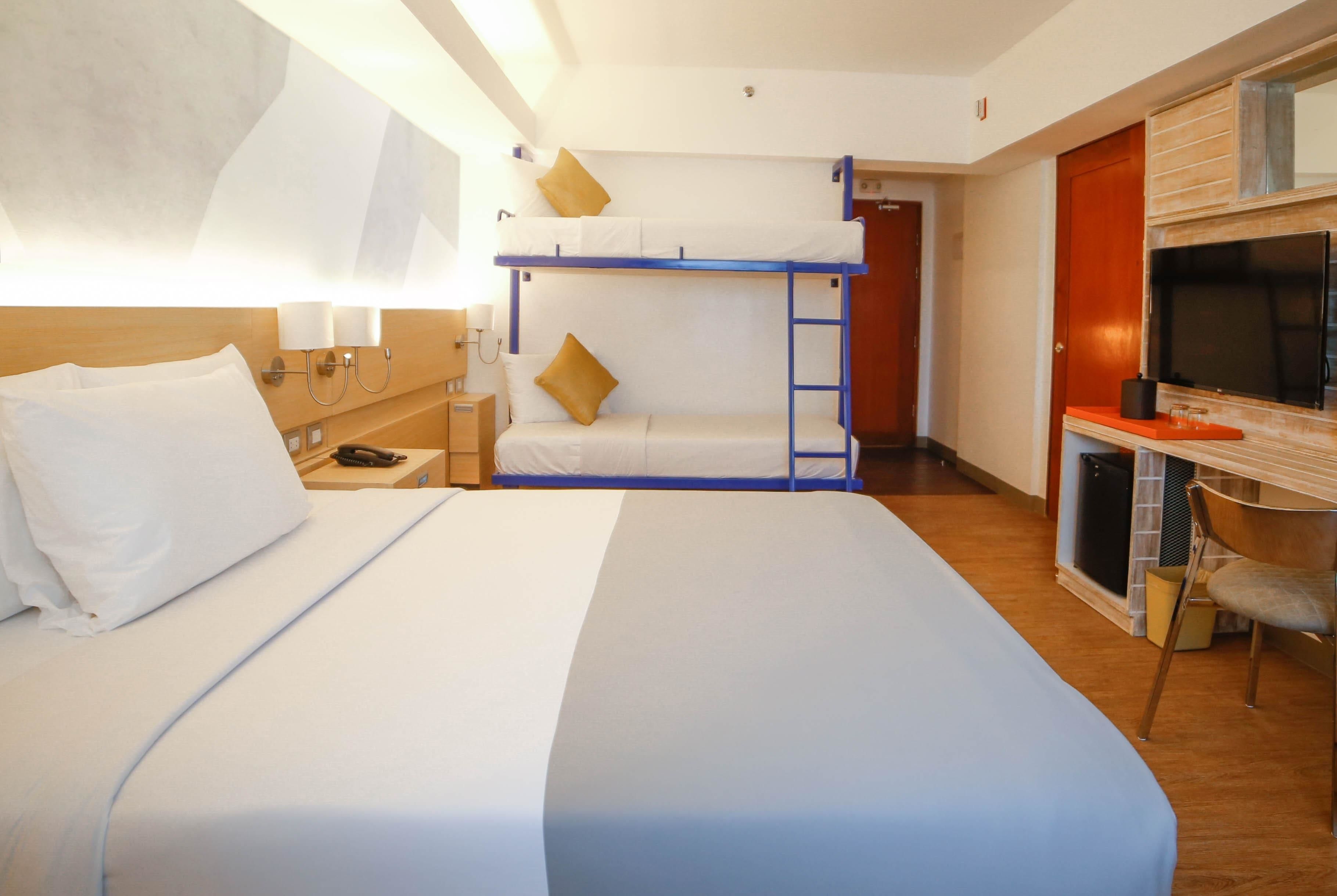A hotel room with a bunk bed, a desk with a TV, and a bedside table with a lamp.