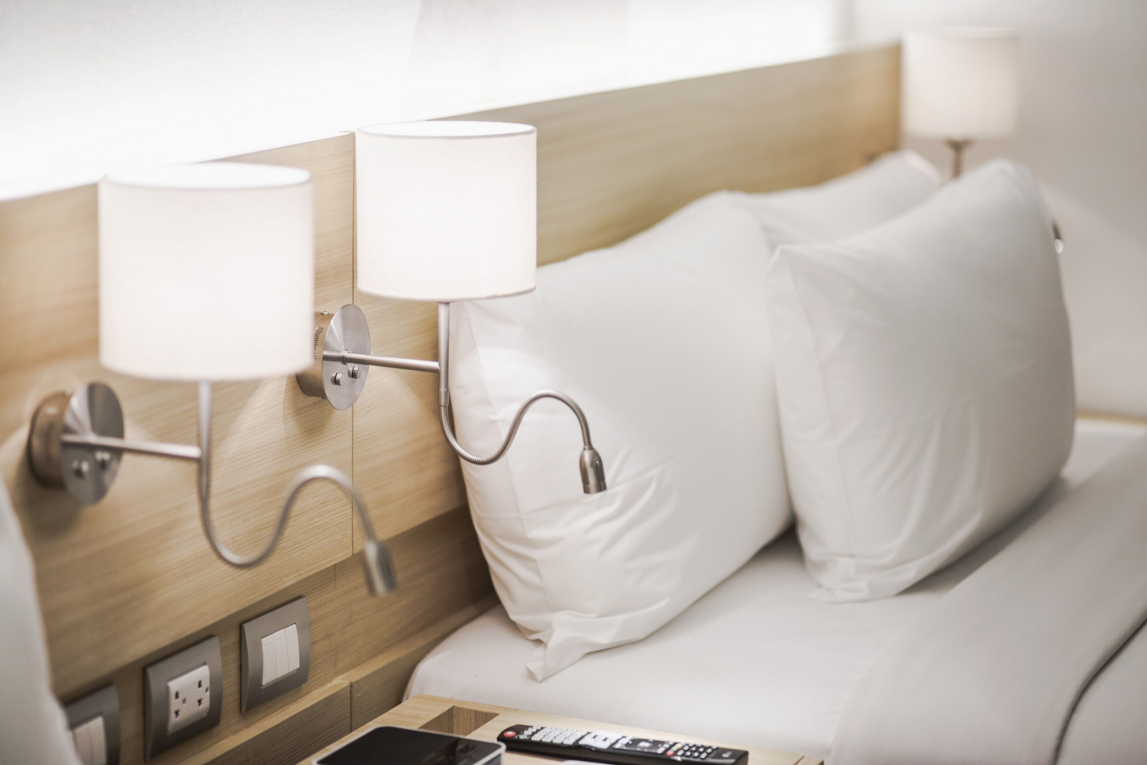 A hotel room with a wooden headboard, two bedside lamps, a remote control, and a smartphone on the nightstand.