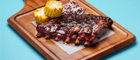 Una tabla de madera con costillas a la parrilla, mazorcas de maíz y ensalada de col.